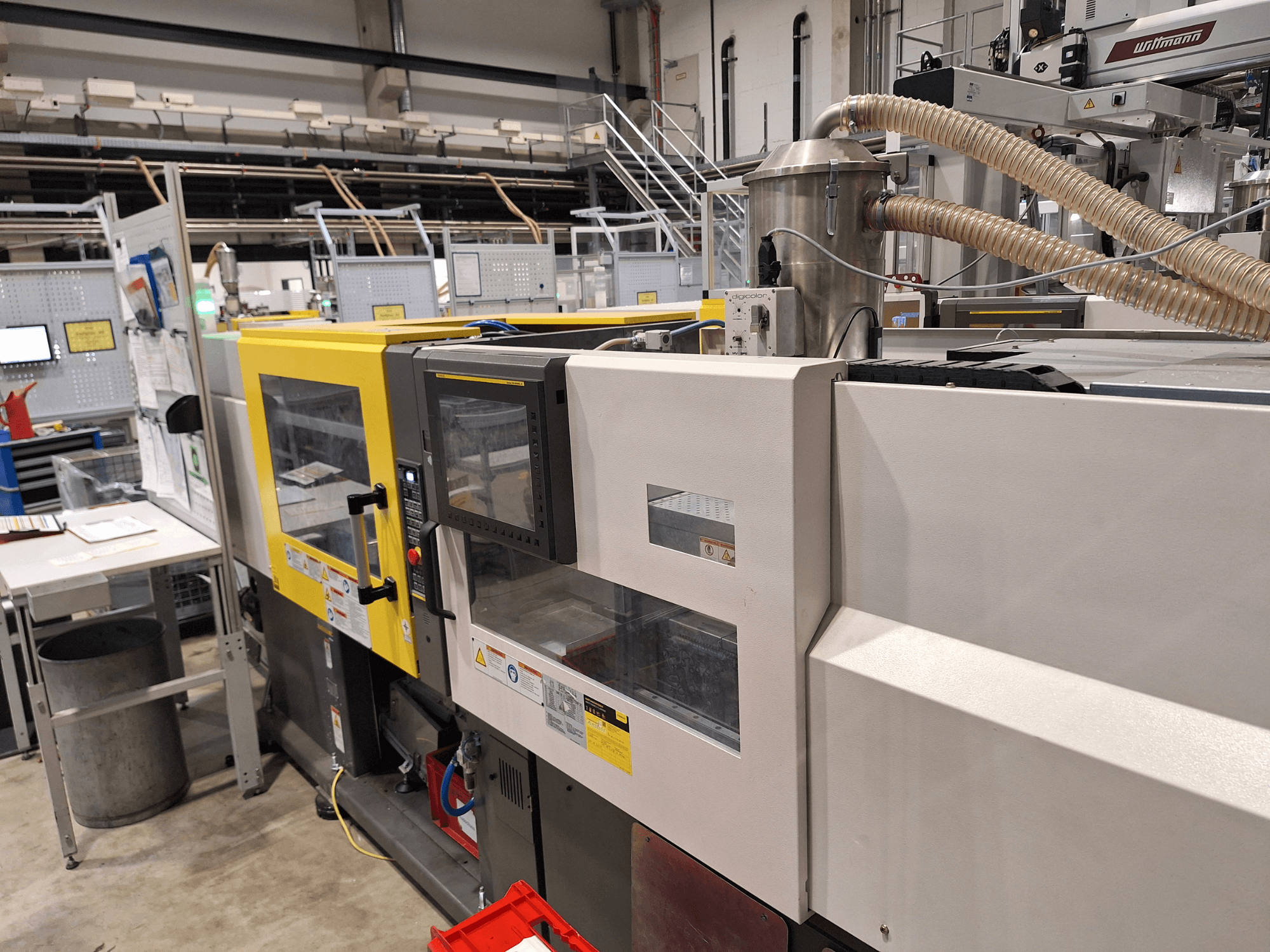 Prednji pogled na višebojni stroj za brizganje plastike, sa žutim i sivim dijelovima, s upravljačkom pločom i ispušnim sustavom.